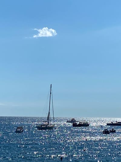 This is a vertical, outdoor, eye-level shot of the Adriatic Sea in Petrovac centar, Montenegro, on a bright, sunny day. The water is a deep blue and reflects the sunlight, creating a sparkly, shimmering effect. In the foreground, the sea is choppy with many bright highlights. Several boats are visible on the water. On the left, a dark-colored, semi-submerged boat is present. To the right of it, a large sailboat with a dark hull and white sails is anchored, its mast reaching high into the sky. Further in the distance, a line of smaller boats, possibly tour boats, are scattered across the water, one with a dark canopy and several passengers. In the far right background, a structure resembling a pier or a breakwater is visible, with what appears to be a person standing on it. A single, small, fluffy white cloud is in the upper left quadrant of the frame against a clear, bright blue sky.