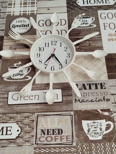 The image shows a close-up of a wall clock with cutlery as hour markers, set against a collage of coffee-themed graphics and textures. The clock face is white with black numbers and hands. Cutlery items such as spoons, forks, and knives are arranged around the clock face to represent the hours. The background features various textures and text, including images of coffee cups, plaid patterns, and words like "HOME," "GOOD FOOD," "TEA TIME," "Green Tea," "LATTE," "Espresso," "Macchiato," and "ALL WE NEED IS COFFEE." A graphic of a teacup with the text "You're my cup of tea" is also visible. The overall impression is of a decorative item for a kitchen or dining area. The image does not contain any information that could indicate its location in Крывасельскі сельскі Савет, Belarus.