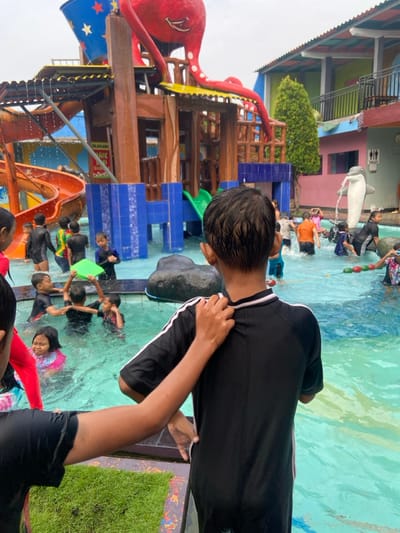 The image shows a waterpark in Kecamatan Tanggulangin, Indonesia. The image was taken at the location (latitude: -7.497, longitude: 112.684). It appears to be a sunny day, and the pool is filled with water. The image captures a boy in the center wearing a black swim suit with white stripes. The boy is being touched on the back by another person, whose arm appears in the image. The boy is looking towards the right side of the image. There are several other children and adults in the waterpark, including a group of children in the water, a group of people standing near the slide, and an adult holding a green rectangular float in the water. The waterpark is decorated with a brightly colored octopus sculpture.