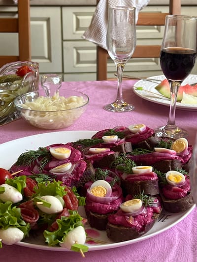 This image depicts a table set with a meal, likely in a home dining area within Mezhdurechenskiy, Russia. A pink tablecloth covers the table, on which several dishes are arranged. In the foreground, a white platter holds numerous open-faced sandwiches topped with a vibrant pink spread, likely beet-based, along with sliced hard-boiled quail eggs, red onion rings, and fresh dill. To the left of the platter, skewers with cherry tomatoes, small white mozzarella balls, and rolled salami slices are presented. Behind these dishes, a clear glass bowl filled with shredded cabbage salad is visible, and further back, a crystal bowl contains pickles and a tomato. Two stemmed glasses are present: one is empty and appears slightly frosted, while the other contains red wine. A white plate with slices of watermelon sits between the glasses. The background reveals a wooden chair and kitchen cabinetry, suggesting an indoor domestic setting. There are no people visible in the image.