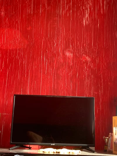 The image displays an indoor scene, likely a room in Troyan, Bulgaria, characterized by a strikingly red wall that occupies most of the left and central portion of the frame. This wall features a textured, distressed finish with numerous fine, horizontal, lighter red and white brushstroke-like lines, giving it a raw or artistic appearance.
To the right, a black, rectangular flat-screen television is prominently positioned. Its screen is dark, reflective, and appears to have a fine layer of dust or smudges. A faint white logo, possibly "LG," is discernible on the bottom right corner of the screen. The TV is supported by a black stand, partially visible beneath its right side.
Above the television and extending towards the top right corner of the image, parts of a shelf or surface are visible. On this surface, a small, golden trophy with a dark base can be seen, its details unreadable. Next to the trophy, a segment of a gift bag or box, featuring a floral pattern and a string handle, is also present.
The scene is static, with no activity taking place. The lighting suggests an indoor setting, but there are no specific cues to indicate the time of day or external weather conditions. The overall impression is that of a decorated interior space with a bold color scheme.