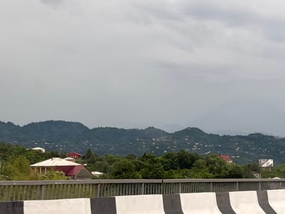This is a landscape photograph taken from an elevated position, likely a bridge or highway overpass, looking towards a mountainous region under a cloudy sky. In the foreground, a concrete barrier with black and white stripes runs horizontally across the bottom of the frame. Beyond the barrier, lush green trees fill the midground, interspersed with several houses. One prominent house in the mid-left features a light-colored exterior and a distinctive red, pitched roof. To its right and slightly further back, another house with a red roof is visible amidst the foliage. Further in the distance, the scene transitions to rolling, densely forested mountains. Scattered across the slopes of these mountains are numerous small, light-colored buildings, indicating a rural or semi-rural settlement. The overall atmosphere is overcast and possibly humid, consistent with a temperate climate. There are no visible people or vehicles. The location is described as Buknari, Georgia.