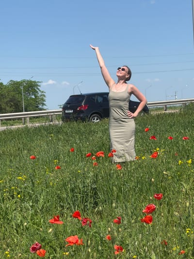 A woman is standing in a field of red and yellow wildflowers next to a black hatchback car on the side of a highway in Novoalexandrovka, Russia. She is wearing a light green dress and sunglasses, with one arm outstretched toward the sky and the other on her hip. The blue sky is visible above, and a line of trees is visible in the background, adjacent to the highway.