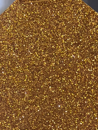 The image is a close-up, overhead shot of a textured surface covered in gold glitter. The glitter appears to be applied to a dark base material, forming a dense, sparkling pattern. The lighting highlights the reflective qualities of the glitter, creating numerous small points of light across the surface. The image is abstract and does not contain any discernible elements of a cityscape, environment, people, or actions. Therefore, it is not possible to provide details about the location in the city of Arcahueja, Spain, as the image does not depict such a setting.