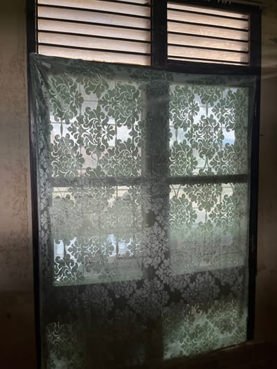A light green, sheer curtain with a damask pattern is hung across a window. The window has a series of horizontal louvers at the top and a grid of glass panes below. The wall to the left of the window appears discolored and stained. The scene is dimly lit, with light filtering through the louvers and the curtain. The image does not contain any people or specific details that would indicate its location in Kota Kuala Simpang, Indonesia.