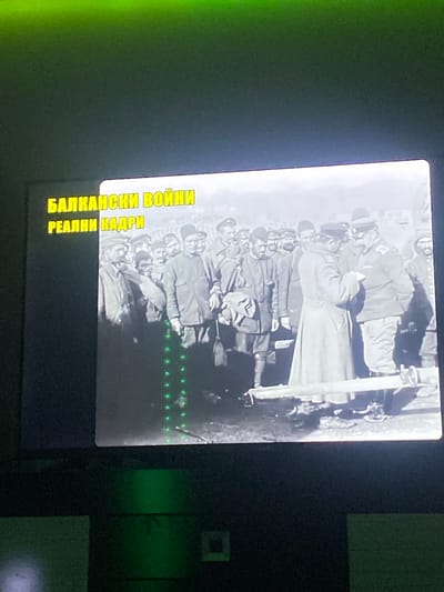 The image displays a black and white photograph, likely a still from a historical film or documentary, projected onto a screen. The photograph depicts a group of men in military uniforms, appearing to be soldiers from an earlier era, possibly the early 20th century. They are gathered in what appears to be an outdoor setting. Some men are looking towards the camera, while others are engaged in conversation or activity. The uniforms suggest a formal or ceremonial occasion.
Overlaying the bottom left corner of the projected image are two lines of yellow text, likely a title or subtitle. The first line reads "БАЛКАНСКИ ВОЙНИ" which translates to "Balkan Wars." The second line reads "РЕАЛНИ КАДРИ," meaning "Real Footage."
Below the projected image, there are prominent green lights forming a vertical line with evenly spaced circular or oval shapes. These lights appear to be an external light source or a projection artifact, not part of the original photograph. The background beyond the screen is dark, with some areas exhibiting a greenish hue, possibly from ambient lighting or the projection system. The overall environment suggests an indoor viewing space where the image is being presented. There is no discernible evidence within the image to confirm or deny a specific location in the city of Benkovski, Bulgaria.