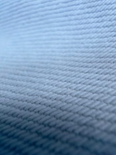 This media file is a close-up, abstract photograph of a textured blue fabric. The fabric appears to be woven, with a pattern of interlocking threads creating a ribbed or corded surface. The lighting creates shadows and highlights that emphasize the texture, giving the fabric a three-dimensional quality. The left side of the image is softly blurred and fades into a lighter blue, suggesting depth and a light source from that direction. The right side of the image is sharper and darker, revealing the intricate weave of the fabric more clearly. There is no discernible subject, activity, or setting beyond the fabric itself. No text is visible. The overall impression is one of detail and texture, with a cool color palette.