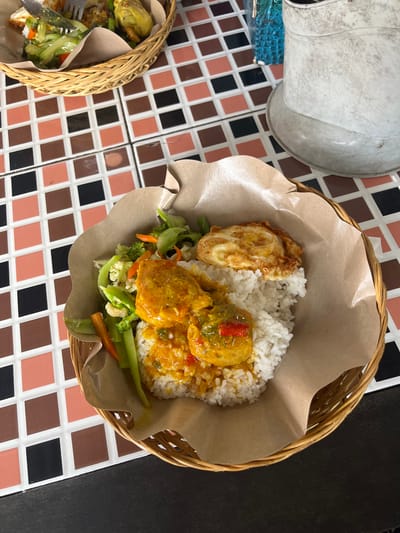 A basket containing a meal is on a tiled table. The meal consists of white rice, a fried egg, vegetables, and meatballs covered in sauce. Another basket with food is visible in the lower left corner. The table has a checkered pattern of brown, black, and peach-colored tiles. The setting is in Kerobokan Kelod, Indonesia.