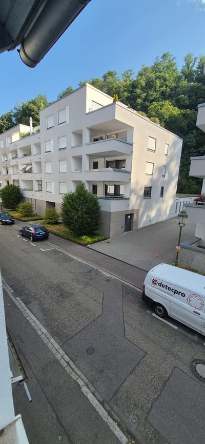 In Baden-Baden, Germany, a modern, multi-story apartment building with white facades and balconies is situated next to a tree-lined hill. A paved street runs in front of the building. Two cars, a dark sedan and a blue Audi, are parked on the side of the street. Further down the street, a white van with the logo "detecpro DIE ORTUNGSPROFIS" is parked. A lamppost stands on the sidewalk. The sky is clear and blue.