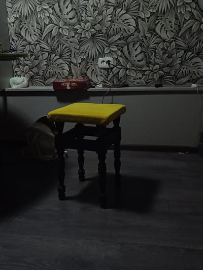 The image depicts an indoor scene with a dark wooden floor and a grey wall with a tropical leaf-pattern wallpaper. In the center of the frame is a dark-colored stool with a bright yellow cushioned seat. To the left of the stool, a light-colored backpack lies on the floor, partially obscured by shadows. On a shelf or ledge behind the stool, a red box with Russian text ("РУССКОЕ ЛОТО") is visible, along with a white plastic bag and a white electrical outlet with a plug. The lighting is dim, casting shadows and contributing to a somewhat moody atmosphere. The specific location in the city of Lermontov, Russia cannot be determined from the image alone.