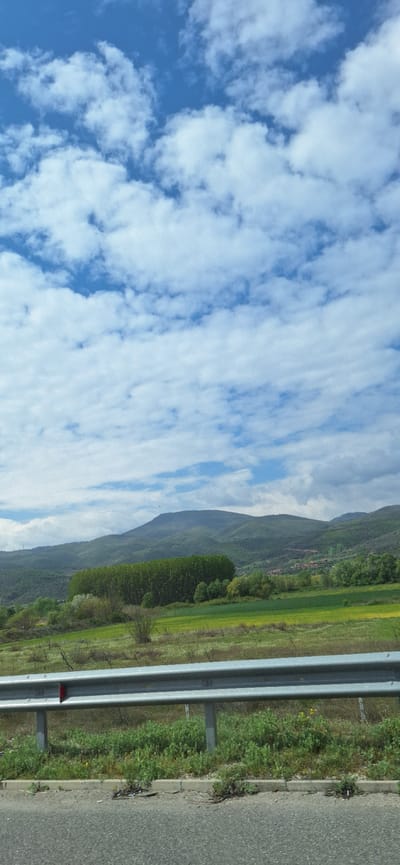 A landscape photograph taken from a moving vehicle depicts a blue sky dotted with fluffy white clouds filling the left side of the frame. The right side shows a roadside environment, including a metal guardrail, a grassy verge with some wildflowers, and a stretch of asphalt road. Beyond the roadside, a green field extends towards a mountainous terrain. The location is Valkovo, Bulgaria, based on the landscape and vegetation. There is no visible human activity.