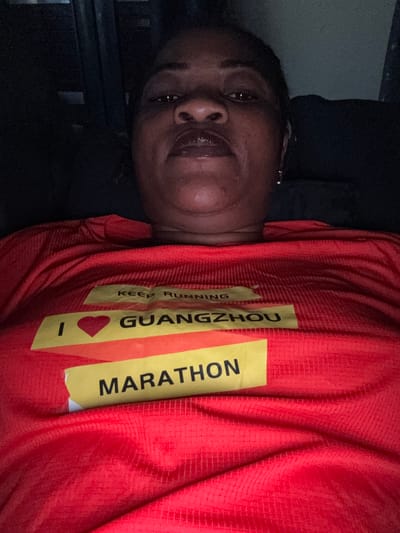 The image shows a close-up of a woman wearing a red t-shirt with yellow text. The woman is lying down, facing upwards, with her head slightly tilted. She has dark skin and appears to be wearing makeup. Her eyes are open and she is looking towards the camera. The t-shirt has text that reads "KEEP RUNNING GUANGZHOU I ❤️ MARATHON". The setting appears to be indoors, possibly a bedroom, given the dark background and what looks like furniture. The lighting is dim, suggesting it might be nighttime or an indoor setting with low light. The woman's expression is neutral. The text on the shirt indicates she is participating in or supporting the Guangzhou Marathon. The provided location context of Ajara, Nigeria, does not have any direct visual cues within the image itself, but suggests the origin or context of the photograph.