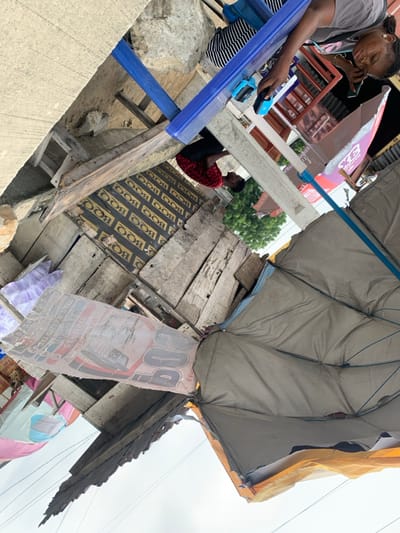 This image, rotated approximately 90 degrees counter-clockwise, depicts an outdoor commercial or market scene in Sangotedo, Nigeria.
In the foreground, a concrete ledge or wall is visible. Below this, a mix of rough wooden planks and exposed dirt constitutes the ground and base of makeshift structures. A prominent, large, beige or tan umbrella with an orange rim, supported by a blue pole, extends into the lower right of the frame, providing shade.
Two individuals are visible near a blue counter or table structure. A woman, wearing a grey short-sleeved top and a black and white striped skirt, stands behind the blue counter. She is holding a dark-colored mobile phone in her left hand and appears to be interacting with a small blue electronic device, likely a POS terminal, also on the counter. To her left, another person, wearing a red patterned short-sleeved top and dark trousers, stands or leans near the counter.
Below the blue counter, a surface of dark wooden planks, repeatedly printed with "BOO" in light text, is present. To the left of this, a large, torn, and faded sign prominently features "BOO" and mentions "WATER," affixed to a wooden structure.
In the background, behind the blue counter, a building structure with concrete pillars is partially visible, along with a red awning or umbrella displaying "3X." Green foliage from trees is seen behind these structures. The sky overhead appears bright and overcast. Faint electrical wires traverse the upper part of the background.
