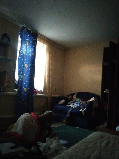 The image depicts a dimly lit bedroom. A blue curtain with white polka dots and cartoon characters hangs over a window. A dark blue couch is positioned in the room, covered with various items of clothing. To the right, a dark wooden shelf unit is visible. In the foreground, a plush toy is lying on the floor. The walls appear to be painted a light brown. The floor is covered with a green rug. The overall impression is of a messy and cluttered living space. The location is specified as a bedroom in Aktobe, Kazakhstan.