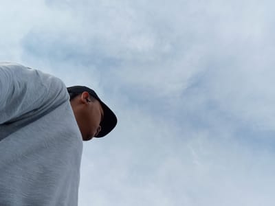 The image shows a person wearing a light-colored, textured t-shirt and a black baseball cap, viewed from a low angle. The person's face is partially obscured as they look downwards. The background is a cloudy sky. No specific location in Kualu, Indonesia, can be determined from this image.