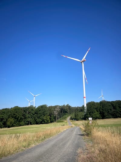 This image depicts a rural landscape with multiple wind turbines under a clear blue sky. A dirt road curves through a grassy field, leading towards a dense line of trees. In the foreground, dry, tall grass lines the right side of the road. Several wind turbines are visible in the background, with the closest one dominating the right side of the frame. The wind turbine in the foreground is white with red accents on its blades and tower. There are no people or animals visible in the scene. The environment appears to be a natural landscape, likely in a rural or semi-rural area, specifically identified as Berfa, Germany.
