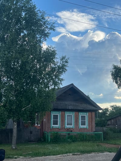 The image shows a traditional wooden house with brick sections in Пигозино, Russia. The house has a dark, steeply pitched roof and windows with decorative green trim. Several trees surround the house, and a green metal fence is visible. The sky is blue with dramatic, large white clouds, suggesting a sunny day with potential for changing weather. Electrical wires are strung across the frame, common in rural settings. The overall scene depicts a quiet, possibly rural, residential area. There is a small plaque with the number '9' on the side of the house. The lighting indicates it is daytime, likely late afternoon or early evening given the soft light and shadows.