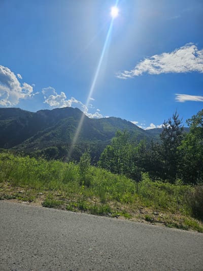 The image depicts a sunny day near Devin, Bulgaria. The scene includes a road running along the right side of the frame. To the left of the road, vegetation grows on the hillside. In the background, there are tree-covered mountains. The sky is blue with scattered white clouds and the sun shining. The overall environment is natural and rural.