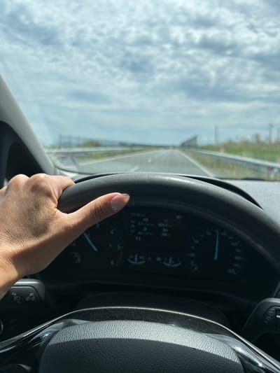 A first-person perspective shot of a person driving a car. The hand holding the steering wheel is in the foreground, and the dashboard with gauges and indicators is visible. The road ahead stretches into the distance, with a bright, cloudy sky above. The image appears to be taken from inside the car, looking forward. The location is stated to be the city of Aheloy, Bulgaria.