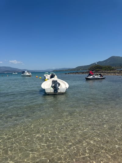 In Lichada, Greece, a clear blue sky stretches over a tranquil seascape. The foreground is dominated by the shallow, crystal-clear waters of the sea, revealing the sandy seabed below. Sunlight dances on the water's surface, creating intricate patterns of light and shadow. Moored in the water are several vessels, including a white motorboat with an outboard motor and a jet ski with two occupants. In the mid-ground, a larger white motorboat is visible, along with two yellow buoys. Further out, a white yacht with several people on board can be seen. The background features a coastline with lush green hills and mountains, bathed in sunlight.