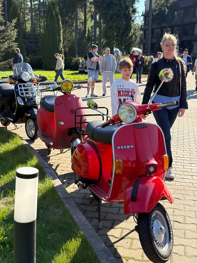 A red Vespa scooter is parked on a brick-paved surface in Repino, Russia, with a boy sitting on it and a woman leaning against it. There are more people and vehicles, including other scooters and cars, in the background. Trees are visible behind the crowd. A black and white pole stands near the scooters on a patch of grass. The day is sunny.