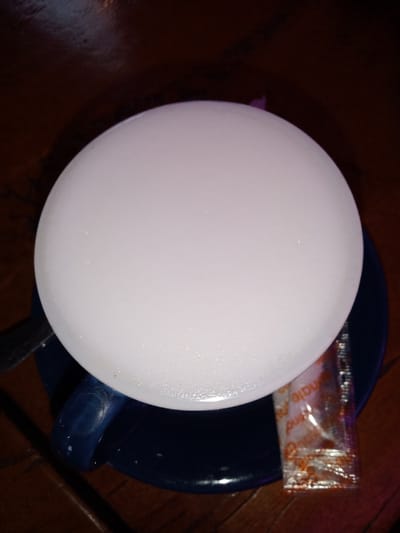 A top-down, slightly angled shot captures a hot beverage served in a dark blue cup on a matching saucer, resting on a dark wooden table. The drink features a thick, bright white, frothy layer, characteristic of a cappuccino or latte, filling the entire visible surface of the cup. To the left, a silver spoon or stirrer is partially visible, resting on the saucer. To the right, a small, transparent rectangular packet, likely containing a condiment like sugar or creamer, lies on the saucer. Part of the text "mate" and "pind" (possibly from "Indomate" or similar branding) is visible on the packet, which has an orange-brown pattern. The lighting is dim, casting a warm glow on the frothy surface while the surroundings remain dark, suggesting an indoor setting such as a cafe or restaurant, likely during the evening or night. No people are visible, and the scene is serene, focused solely on the prepared drink.
