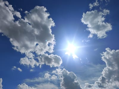 A bright, sunny day is depicted in this image of the sky. Puffy white clouds are scattered across a vibrant blue sky. The sun is shining intensely, creating a lens flare effect and casting rays of light. The lower right corner of the image displays the text "17.06.2026 15:05", indicating the date and time the photo was taken. The scene suggests fair weather and a pleasant afternoon. The location context is Үштөбе, Kazakhstan.