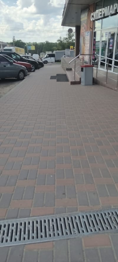 This is a vertical, medium-shot, exterior, daytime photograph taken on a cloudy day in Valky, Ukraine. The scene depicts a street in front of a supermarket. The foreground is dominated by a tiled sidewalk with a metal drainage grate running diagonally across it. In the background, several cars are parked along the side of the road. Beyond the cars, there is a row of trees and a building that appears to be the supermarket, with large glass windows. The text "СУПЕРМАРК" (SUPERMAR) is visible on the front of the building, indicating it is a supermarket. The sky is filled with white clouds. The overall impression is a typical street scene in a Ukrainian town.