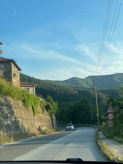 This media file, oriented vertically, captures a road winding through a lush, mountainous landscape. The scene appears to be a bright, sunny day, possibly late morning or early afternoon, with a clear blue sky dotted with wispy white clouds.
The foreground features a rough concrete embankment covered in dense green vegetation. Part of a rustic stone building with a tiled roof is visible on the left, suggesting a rural or older settlement. Power lines stretch across the upper portion of the frame, supported by a wooden utility pole.
The road itself curves to the right, with a white car driving on it. A series of traffic signs are mounted on poles alongside the road, indicating a sharp curve to the left, a "no stopping" zone, and a speed limit of 30. The road surface appears wet in places, reflecting the sunlight.
In the background, a dense forest blankets the steep slopes of a mountain range, its contours softened by distance and the hazy atmosphere. The overall impression is one of a scenic, somewhat remote location, likely Taran, Bulgaria, characterized by natural beauty and a peaceful, everyday atmosphere. There is no discernible emotional content or specific event taking place beyond normal travel.