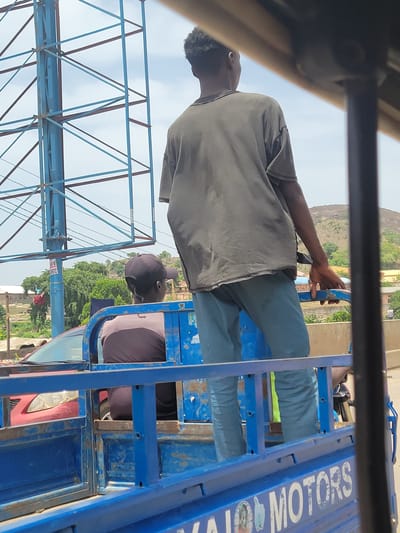 From within a vehicle, a man in a gray shirt and teal pants drives a blue tricycle through Suleja, Nigeria. Another man, wearing a black cap and brown shirt, rides in the back. The vehicle is branded with "JAIJO MOTORS." In the background, a hill and a billboard structure can be seen. Other vehicles are visible in the periphery.