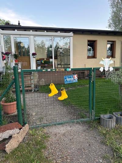 The image depicts the exterior of a residential property in Wustermark, Germany, focusing on a green metal gate leading into a yard. The gate is partially open, revealing a lawn and the back of a building with windows and an extension. Two yellow rubber boots are hanging from the fence. A sign on the gate reads "Es gibt sooo viele Hunde auf der Welt und ausgerechnet hier lebt der Beste" (There are so many dogs in the world, and by chance, the best one lives here). A white, cartoonish cow figure is attached to the top right of the gate. In the foreground, there are some discarded bricks and gravel. The sky is visible in the background, with some trees and clouds. The overall scene suggests a domestic outdoor setting.