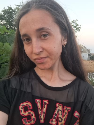 A selfie portrait of a young woman with long, dark brown hair, freckled skin, and brown eyes. She is wearing a black t-shirt with mesh paneling across the chest and leopard print letters that read "SV". She is outdoors with greenery and trees visible behind her. In the background, a house with a white facade and a gray roof can be seen. The overall setting appears to be rural or suburban. The image was captured in Vypasne, Ukraine.