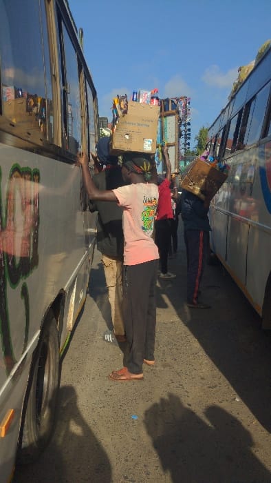 Three people are carrying items on their heads. They are standing close to parked buses in what appears to be an open area or market. The person closest to the camera wears a light pink shirt with an illustration, dark pants, and red sandals. The second person is wearing a red shirt and dark pants. The third is wearing a dark long-sleeved shirt and dark pants with a light colored stripe. The items on their heads appear to be boxes or containers.