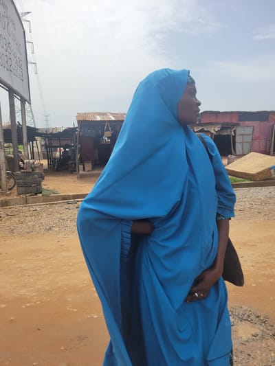 A woman in a blue garment stands on a dirt road in Kwamba, Nigeria. Background: shacks and utility poles.