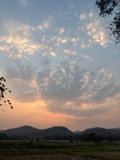 The scene depicts an outdoor landscape in Ban Huai Sai, Thailand, at sunset. The sky is largely covered with scattered altocumulus clouds, presenting a gradient from light blue at the zenith to vibrant orange and pink hues along the horizon. The undersides of many clouds are illuminated with warm, golden light. Dark, silhouetted tree branches with visible leaves partially frame the image from the upper right and mid-left. In the midground, a range of dark, forested mountains or hills spans the width of the frame, their contours obscured by low light. On the peak of the leftmost prominent hill, a faint, lighter structure is discernible. Below the mountains, the environment transitions to a flat expanse of what appears to be dry, harvested agricultural land, with a darker strip of vegetation in the immediate foreground. Several thin, dark vertical structures, resembling utility poles or fence posts, are visible between the field and the mountain range, with faint horizontal lines suggesting wires or fencing. No people, subjects, or interactions are visible.