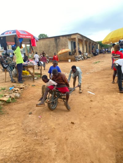 The image depicts an active outdoor market or street scene in Riyom, Nigeria, characterized by unpaved, sandy ground under an overcast sky. The overall impression is one of daily life and commerce in a rural or semi-urban setting.

In the immediate foreground, two young children are engaged with a small, metal-framed bicycle or tricycle. One child, wearing a red and brown patterned shirt and red shorts, is seated on it, while another child in a blue shirt appears to be interacting with or assisting them. Slightly further back, a man in a light-colored long-sleeved shirt is pushing a small, two-wheeled handcart or wheelbarrow across the dirt path.

Along the right side of the scene, several market stalls are visible, constructed from wooden tables. Various goods, including clothing, are displayed on these stalls. Numerous individuals, including men and children, are present around these stalls, either browsing, selling, or observing. Above one of these stalls, a red canopy is visible, bearing the text "YIELDS MORE" and "3X".

In the background, on the left, stands a multi-story building with a light-colored facade and visible columns, suggesting a permanent structure. Motorcycles are parked near this building. A yellow canopy is also present in the distance on the left. The scene is bustling with people engaged in various activities, indicative of a lively market environment during daytime.