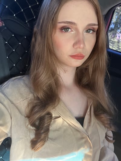 A young woman with long, wavy, light brown hair is shown from the chest up, looking forward. She is seated in the passenger seat of a vehicle, as indicated by the car seat behind her and the window with a blurred outdoor scene to her right. She is wearing a light beige button-up shirt. Her makeup includes eyeshadow and lipstick. The scene appears to be captured during daylight hours. There is no discernible evidence to suggest this image was taken in the city of Новая Отрадовка, Russia.