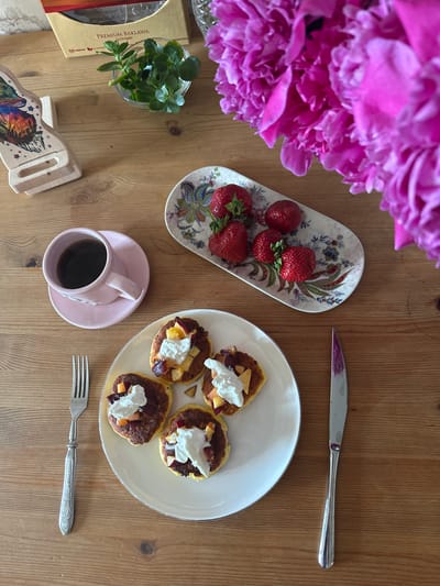 This is a top-down, medium shot of a breakfast spread on a wooden table. In the upper left is a box of "PREMIUM BAKLAWA". To the right of the box is a small glass bowl with green herbs. To the right of the herbs are vibrant pink peonies. Below the baklava box is a wooden phone stand with a colorful fox design. In the center left, a pink teacup and saucer holds black coffee. To the right of the teacup is a small oval plate with several strawberries. In the center right, a white plate holds four small pancakes topped with whipped cream and fruit. A silver fork rests to the left of the white plate, and a silver knife with a purple stained blade rests to the right. The table surface is made of light brown wood planks. No people are visible in the image. This scene is not specifically located in Vladimir, Russia.