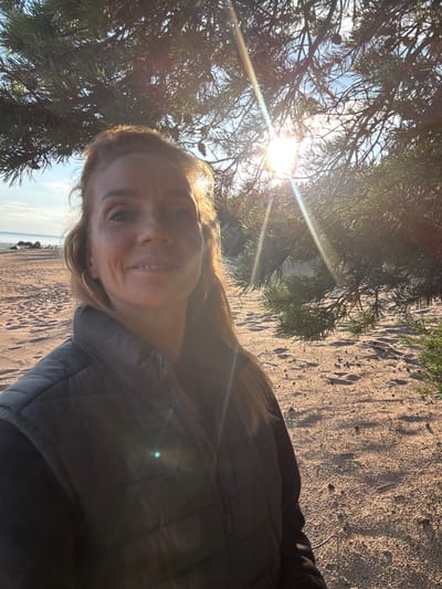 A woman is taking a selfie on a sandy beach with pine trees and the sea in the background. The sun is shining brightly through the pine needles, creating lens flares in the image. The woman is wearing a black quilted vest over a black garment and large hoop earrings. The beach is covered in sand and pine cones, with footprints visible in the sand. In the background, the sea is calm and the sky is blue. The location is the shore of the Gulf of Finland, near Shepelevo, Russia, an area known for its sandy beaches and pine forests.