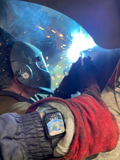 The image depicts a person welding in what appears to be an industrial or workshop setting. The primary subject is a welder wearing protective gear, including a welding mask with the name "HASSAN" written on it in white marker, and heavy-duty gloves. One hand, covered by a red and grey glove, is positioned near the welding torch, emitting a bright, intense light and sparks. The other hand, also gloved, is visible in the foreground, wearing a smartwatch. The watch face displays a globe image, the time "14:51," the date "СР. 17" (likely Wednesday, 17th), and some Cyrillic text which reads "ВОСНОВНОМ СОТЕ". Blue smoke or fumes from the welding process are also visible. The lighting is dominated by the bright glare of the welding arc, creating a high-contrast scene with deep shadows. The overall impression is one of focused, industrial work. The location context provided is Tomba, Montenegro.