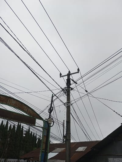This image presents an overhead street scene in Ambakirano, Madagascar, under an overcast grey sky. A prominent concrete utility pole, positioned slightly right of the frame's vertical midline, serves as a central hub for a dense network of black electrical and communication cables. The pole features multiple cross arms with insulators at its upper section, with a substantial cluster of wires, conduits, and equipment affixed below. Numerous black wires, varying in thickness, crisscross the entire upper frame, connecting to the main pole and a secondary, thinner pole or support structure to its left, which also bears a small white junction box. In the lower right quadrant, a section of a building with a heavily rusted, corrugated metal roof is visible, with a darker, indistinct roofline adjacent to it. To the left, a row of tall, dark green, conical evergreen trees is partially visible, appearing behind a weathered, arched metal structure with rust and faded green paint in the lower-left foreground. No individuals are present, and no discernible actions or interactions are depicted within the scene.