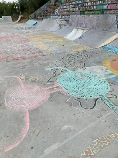 This media file depicts a concrete skatepark in Badalona, Spain, adorned with colorful chalk drawings. The scene is captured from a slightly elevated, oblique angle, giving a sense of the park's scale and the expansive nature of the artwork. The concrete surfaces, including ramps and ledges, are covered with a vibrant array of drawings, suggesting a recent community art event or ongoing artistic expression within the skatepark.

The subjects of the chalk art are diverse, featuring abstract patterns, stylized figures, and what appear to be playful, cartoon-like creatures. One prominent drawing is a large, turquoise, circular shape filled with black dots, possibly representing a stylized insect or a decorative motif. Another striking image is a pink spiral, reminiscent of a candy or a stylized eye. The drawings are rendered in chalk, which gives them a somewhat faded and textured appearance, blending with the rough concrete surface.

The activity taking place is artistic creation or appreciation, as evidenced by the chalk art covering the park. The time of day appears to be daytime, with natural light illuminating the scene. The weather is likely fair, as the chalk drawings are exposed to the elements. There are no people immediately visible in the frame, but the presence of the artwork implies a community that uses and contributes to the space.

Notable details include the vibrant graffiti on the stairs to the left of the frame, which contrasts with the chalk art on the ground. This suggests multiple forms of artistic expression within the same location. The concrete structures of the skatepark are clearly visible, indicating a functional space designed for skateboarding and other similar activities. The overall emotion conveyed is one of creativity, playfulness, and a sense of community engagement with public space. No distinct text is legible in the chalk drawings, though some of the graffiti on the stairs may contain writing, it is not clear enough to read.