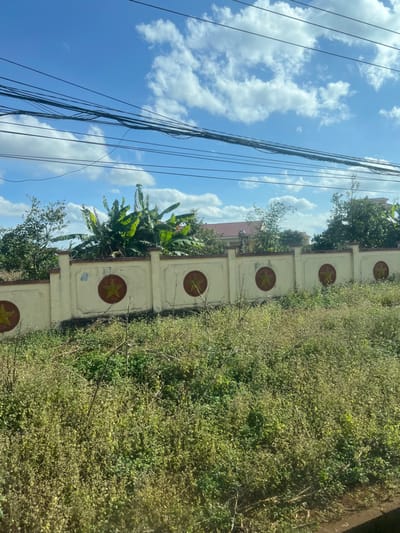 An outdoor scene in Trung Hồ, Vietnam, depicts a concrete wall running horizontally across the mid-ground, featuring five visible circular emblems containing a yellow five-pointed star on a reddish-brown background, spaced along its length. The wall is light beige, exhibiting signs of age and discoloration. Behind the wall, dense green foliage, including banana trees, partially obscures a building with a reddish-brown tiled roof and light-colored walls. The foreground consists of an unkempt patch of overgrown green and brown vegetation. Above the wall and foliage, the sky is clear blue with scattered white cumulus clouds. Multiple black electrical or utility lines are strung horizontally across the upper portion of the sky. No individuals, animals, or active interactions are present in the frame.