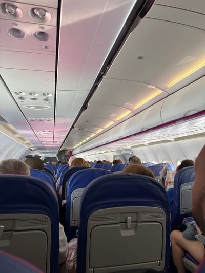 The image shows the interior of an airplane cabin, viewed from a passenger's perspective. Several rows of blue airplane seats are visible, occupied by passengers. The aisle runs down the center of the cabin, illuminated by overhead lights. The scene appears to be inside a commercial airplane during a flight, suggested by the rows of seats, overhead compartments, and the visible text on the seats. The lighting in the cabin is bright and artificial, casting a warm glow on the interior. Some passengers have their heads resting on the seatbacks or are looking down. Text on the seatbacks provides safety instructions in multiple languages, including English, Bulgarian, and Ukrainian, referring to life vests and seat belts. The overall atmosphere appears calm and ordinary, typical of a flight. The location context is Aksakovo, Bulgaria, which implies this is likely a flight arriving at or departing from Bulgaria. There are no clear indicators of time of day or weather conditions from within the cabin.