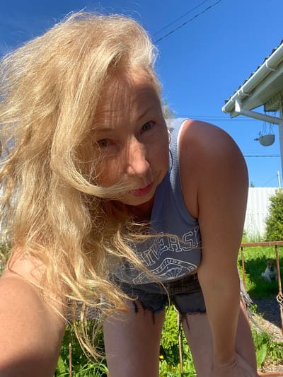 The image shows a woman with long blonde hair, looking at the camera. She is wearing a blue tank top and denim shorts. The photo is taken outdoors, with a clear blue sky in the background. There is a white building with a green roof and gutters on the left side of the frame. A metal fence is visible on the right, with green foliage behind it. A small dog is sitting behind the fence. The lighting suggests it is daytime, likely sunny. The overall mood appears casual and relaxed. Visible text on the woman's tank top includes "HUDE" and "DAY". The location context provided is Bol'shoe Zaborod'e, Russia.