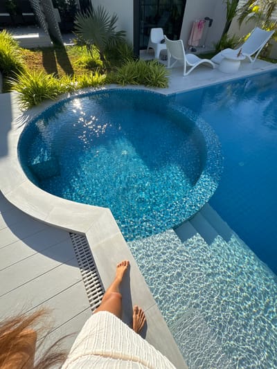 A high-angle, close-up shot captures a portion of a pristine swimming pool area, bathed in bright sunlight, suggesting a warm and clear day. The pool itself is a vibrant blue, with a section tiled in smaller, iridescent blue mosaic tiles forming a circular inset. Steps, also tiled in the smaller mosaic, lead down into the pool. The water reflects the sunlight, creating a shimmering, dappled effect on the pool floor and steps.
To the right of the pool, a person's legs and feet are visible, resting on a light gray wooden deck. They are wearing a white, textured garment. The deck has a modern, slatted drain grate along its edge.
To the left of the pool, a modern villa with white furniture is partially visible. White lounge chairs and a small table are arranged on a white tiled patio. In the background, lush green tropical foliage, including a palm tree, adds to the serene and luxurious setting. The shadows cast by the foliage and the villa indicate it is likely late morning or early afternoon. The overall mood is one of relaxation and summer leisure. No text is visible in the image. The location context is Baan Tai, Thailand.