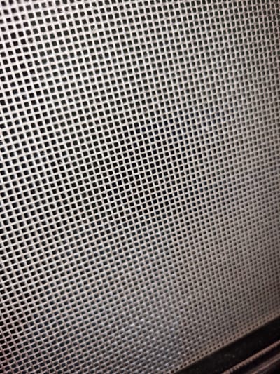 The image shows a close-up view of a window screen. The screen is composed of a tightly woven mesh of thin, light-colored strands forming a grid of small, square openings. Part of the screen's black frame is visible in the upper right corner of the image. The overall setting is indistinct, and the location, Santa Cruz de la Sierra, Bolivia, is not discernible from the image alone. There are no people, subjects, objects, actions, or interactions depicted other than the screen itself.