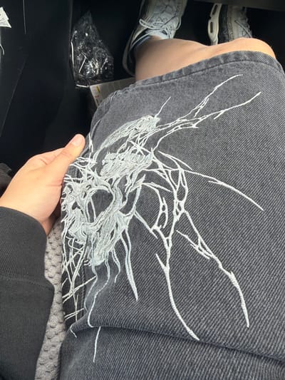A close-up, eye-level, over-the-shoulder shot shows a person's hand resting on their knee, which is covered by dark denim shorts with an intricate white embroidered design. The embroidery appears abstract, resembling tangled branches or veins. The person is wearing a black long-sleeved shirt and a grey cushioned seat is visible. In the background, partially obscured, are a person's legs wearing athletic shoes and a black plastic bag. The scene is indoors, likely in a vehicle, given the limited background detail and the presence of the seat. The location is identified as Beaverton, United States of America.