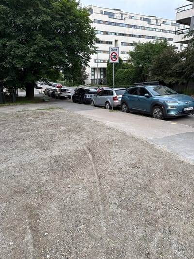 This image shows a street in Oslo, Norway, with several cars parked along the side of a building. The building is modern and features many windows. A speed limit sign is visible, indicating a 30 km/h zone. Trees and greenery are present, suggesting an urban park or residential area. The ground is a mix of gravel and concrete. The lighting suggests it is daytime, possibly overcast.