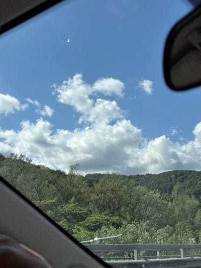 The image captures a view through a car window, looking out at a bright blue sky filled with fluffy white clouds. In the upper portion of the sky, two small, bright white lights are visible, possibly lens flares or reflections. Below the sky, a dense forest covers rolling hills. A grey metal guardrail is visible in the foreground, separating the road from the embankment. The lower left corner of the image shows a portion of the car's interior, including a person's arm and hand. The scene appears to be in a rural or suburban area, given the natural landscape. There is no indication of the specific location within Adlerskiy, Russia, as no landmarks or identifying features are present.